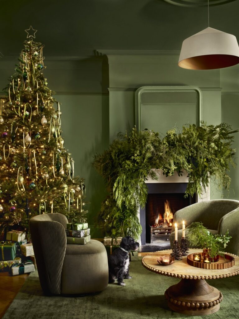 Green living room with armchairs, large foliage festive mantle display above a lit fireplace and Christmas tree with ribbons.